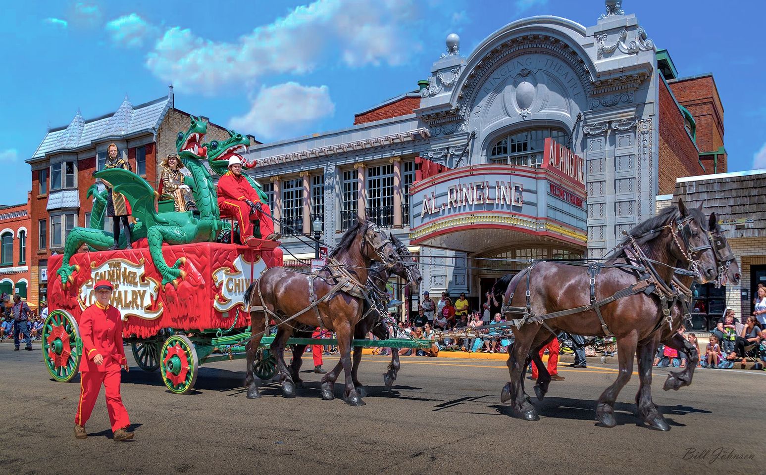 A red carriage wagon with a large green dragon sprawled over the top rides along a street in Baraboo Wisconsin pulled by a team of 4 horses with Circus World Staff wearing read guiding it a long as part of the Baraboo's Big Top Parade Event